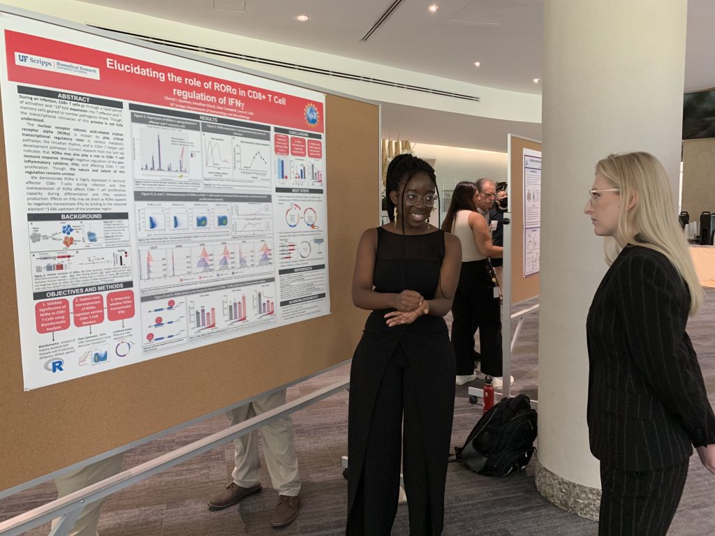 A woman stands next to her research poster titled 'Elucidating the role of RORα in CD8+ T Cell regulation of IFNγ' from UF Scripps Biomedical Research, speaking with another woman The poster contains multiple charts, graphs, and data visualizations on a red and white background.